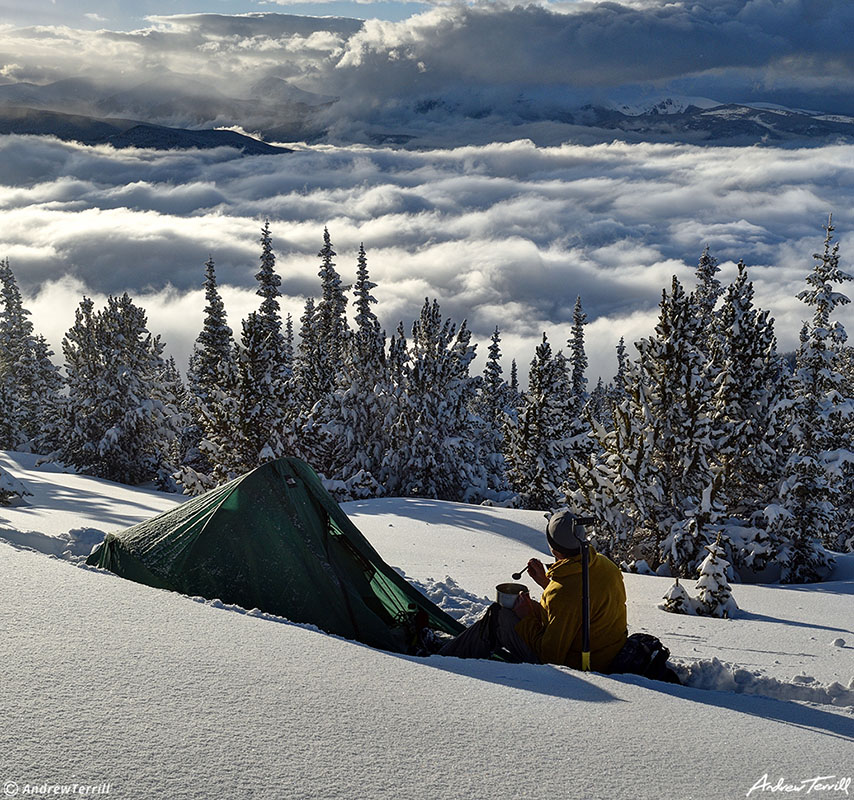 winter wild camping with tent and figure eating dinner looking at view in snow with clearing storm clouds over rocky mountains in colorado